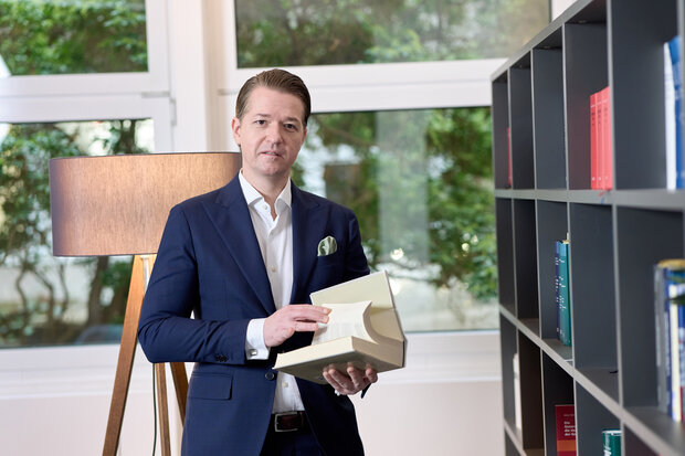 Ein Mann, im dunkelblauen Jackett mit einem aufgeschlagenen Buch.  Querformat.