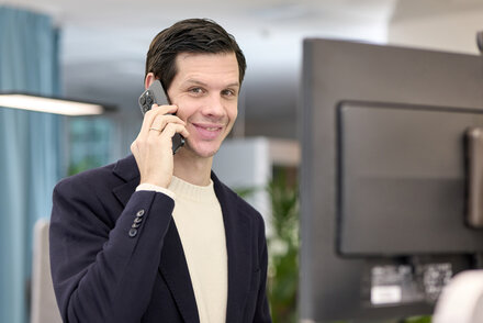 Mann im dunkelblauen Jackett führt ein Telefonat am Schreibtisch, Querformat